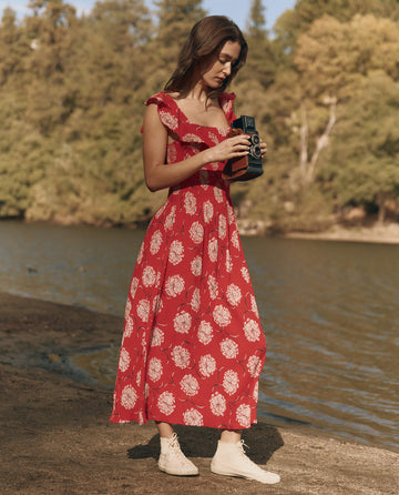 A woman in THE GREAT. SU25 Havana Dress in True Red Dancing Floral stands by a lake, gazing at a vintage camera amid trees—a perfect moment of effortless, elegant fashion.