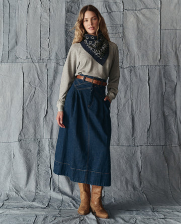 A woman stands against a textured gray backdrop, wearing a gray long-sleeve top, belted high-waisted denim skirt, tan cowboy boots, and The Bandana Kerchief in Navy from THE GREAT. H25 COLLECTION.