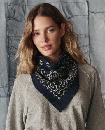 A woman with wavy blonde hair wears a grey long-sleeve top and THE GREAT. H25 COLLECTION The Bandana Kerchief in Navy, standing before a textured grey background.