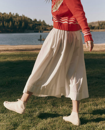 A woman wearing THE GREAT. SU25's The Viola Skirt in white walks on grass by a lake, with trees and hills behind her, capturing the essence of women's fashion.