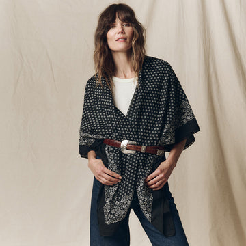 Woman wearing a patterned black kimono-style jacket over a white shirt, blue jeans, and a brown belt, standing against a beige backdrop.