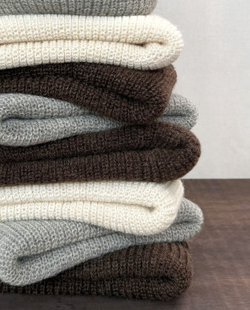 A brown Natural Alpaca Beanie by THE GREAT. HOL 24 BIRKENSTOCK, crafted from merino alpaca blend and undyed yarn, rests neatly folded on a dark wooden surface against a plain background.