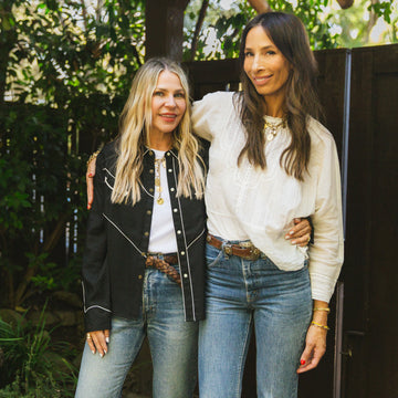 Two women standing side by side outdoors, both wearing jeans and casual tops, smiling at the camera with one arm around each other.
