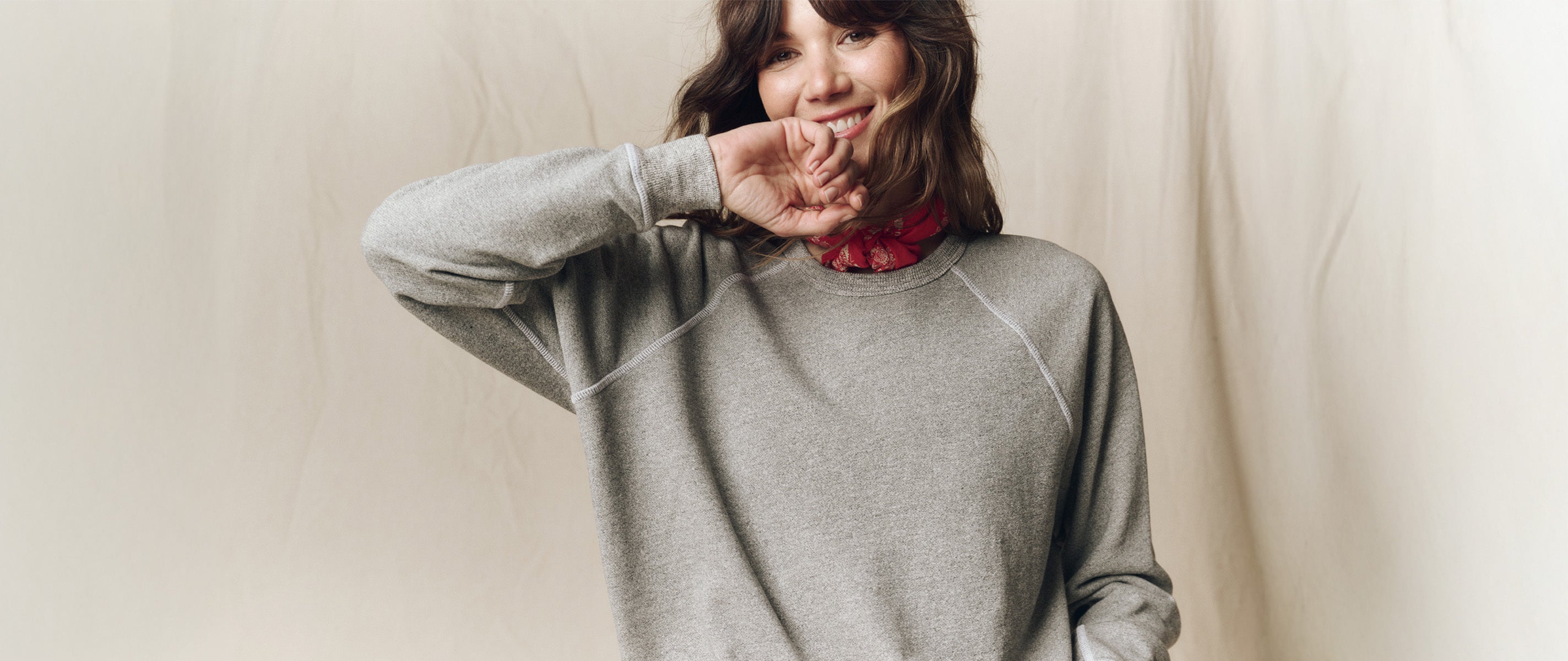 A person wearing a gray sweatshirt and red bandana stands in front of a beige background, raising one hand near their face and smiling.
