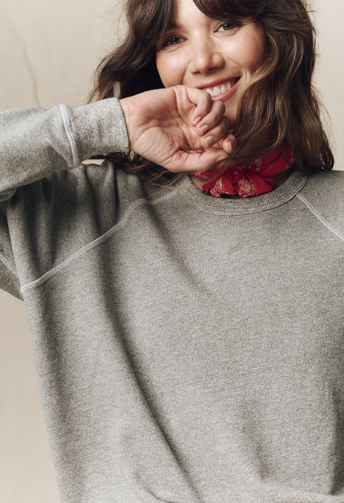 Woman wearing a grey sweatshirt and red patterned scarf, smiling with her hand near her face against a neutral background.