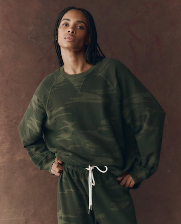 A person wears THE GREAT. F25 D1 The Slouch Sweatshirt. Novelty -- Forest Camo, paired with matching sweatpants featuring a white drawstring, and stands against a brown backdrop.