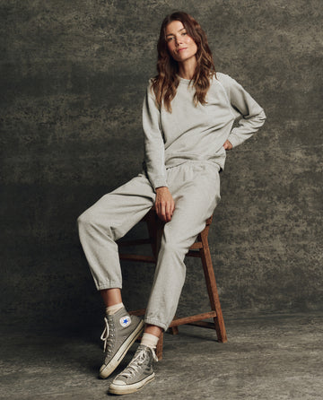 A woman sits on a wooden stool against a textured gray background, wearing THE GREAT. H25 CORE FLEECEBACK The Fleece Stadium Sweatpant in Heather Grey, a matching sweatshirt, and gray Converse sneakers.