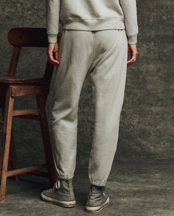 Wearing THE GREAT. H25 CORE FLEECEBACK The Fleece Stadium Sweatpant in Heather Grey, a person stands by a wooden chair, facing away, set against a textured grey background.