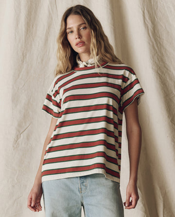 A woman stands before a beige fabric backdrop wearing THE GREAT. F25 D2 Boxy Crew in Cream Bridle Stripe with light blue jeans.