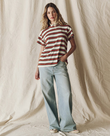 Standing in front of a cream fabric backdrop, the person wears THE GREAT. F25 D2 Boxy Crew Novelty in Cream Bridle Stripe, light blue wide-leg jeans, and sneakers with one hand in their pocket.