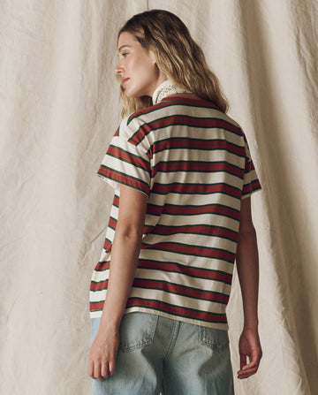 A woman faces away from the camera, wearing THE GREAT. F25 D2's The Boxy Crew. Novelty in Cream Bridle Stripe, paired with light blue jeans, standing before a beige fabric backdrop.