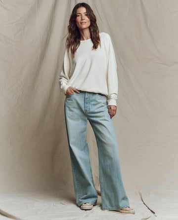 A woman stands before a beige backdrop wearing THE GREAT. CORE KNITS The College Sweatshirt in Solid Washed White—an oversized 100% cotton top—paired with light blue wide-leg jeans and white sneakers.