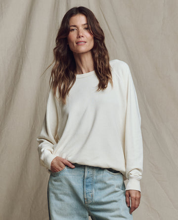 A woman with wavy brown hair wears THE GREAT. CORE KNITS The College Sweatshirt in Washed White with light blue jeans, standing before a beige fabric backdrop.