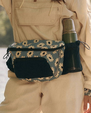 A person in a beige outfit embodies adventure with THE GREAT. SP25 TGO's "The Adventure Hip Pack - Falling Daisy Multi," featuring a floral pattern and a green thermos in its side pocket.