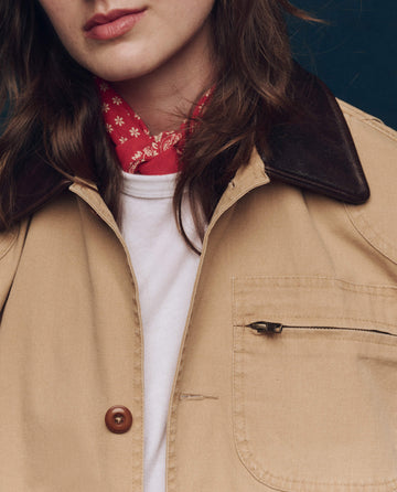 The person wears THE GREAT. SP25 COLLECTORS' Barn Jacket in tan with a leather collar and red bandana, featuring a button front and zippered chest pocket, over a crisp white shirt for a stylish look.
