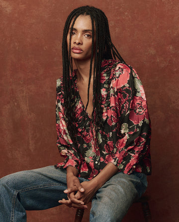 A person with long braided hair in The Symphony Top -- Charming Rose by THE GREAT. HOL 24 and jeans sits gracefully on a chair against a brown backdrop.