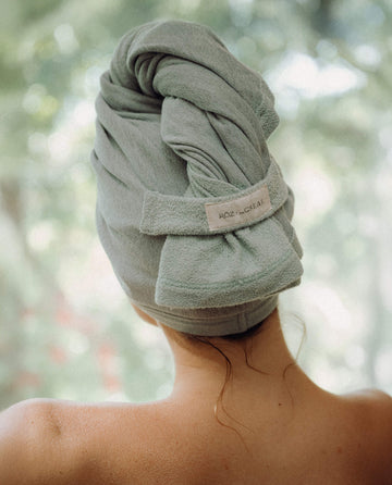 A person stands with their back to the camera, hair wrapped in THE GREAT. PF25 ROZ RŌZ + The GREAT The Sweatshirt Hair Towel – Seaglass; the towel is designed for quick drying and frizz reduction.