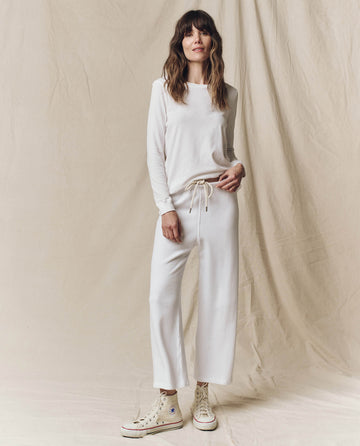 A woman stands against a beige fabric backdrop wearing THE GREAT. F25 CORE KNITS The Long Sleeve Slim Tee in Solid True White, matching drawstring pants, and off-white high-top sneakers.