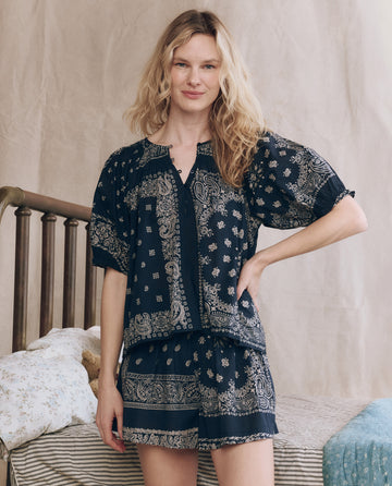 A woman stands indoors in front of a bed with floral bedding, wearing THE GREAT. F25 SLEEP's The Smocked Sleep Top in Bandana Patchwork print and matching shorts, with one hand on her hip.