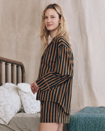 A woman wearing THE GREAT. F25 SLEEP "The Pajama Shirt." in Club Stripe stands beside a bed with white floral pillows and a green coverlet, facing slightly to the side.