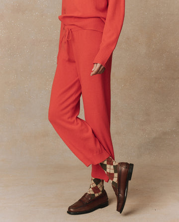 A person dressed in an Ember set from THE GREAT. HOL 24 CASHMERE, featuring a red cashmere sweater and The Cashmere Lantern Pant, stands against a beige background, their outfit completed with checkered socks and brown loafers.