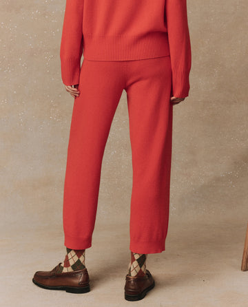 A person stands against a beige background, wearing a red cashmere sweater and The Cashmere Lantern Pant in Ember from THE GREAT. HOL 24 CASHMERE, with patterned socks peeking above brown shoes, facing away from the camera.