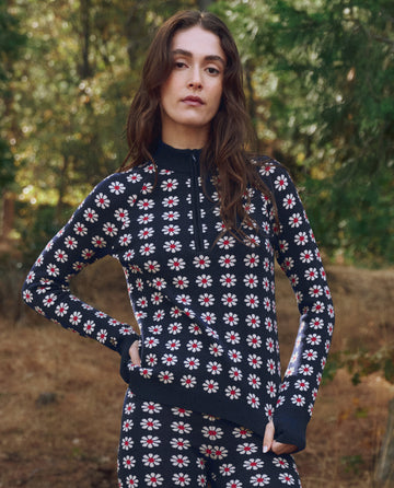 A woman stands outdoors in THE GREAT. F25 TGO’s Merino Quarter Zip in Frosted Daisy, featuring a red, white, and black floral pattern—an ideal slopeside companion amid trees and foliage.