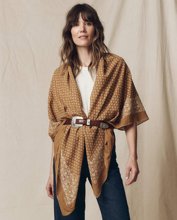 A person elegantly stands against a beige background wearing THE GREAT. SP25 D2's The Big Bandana in Marigold, belted over a white shirt with dark jeans.