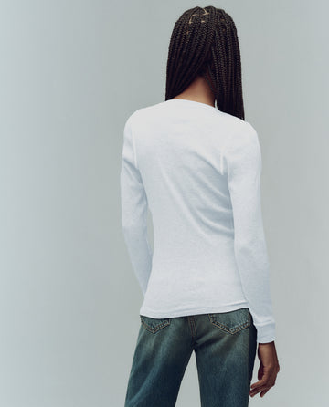 A person with long braided hair stands with their back to the camera, wearing THE GREAT. F25 BABY RIB’s The Baby Rib Henley in True White and blue jeans against a plain light background.