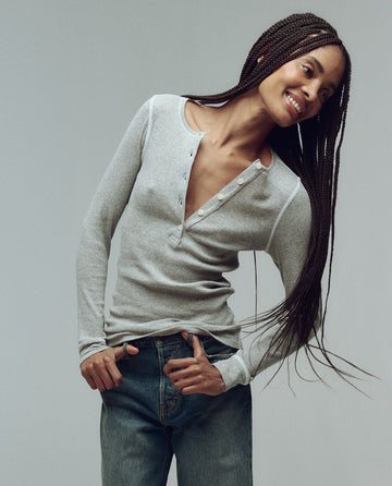 A person with long braided hair, wearing THE GREAT. F25 BABY RIB The Baby Rib Henley in Heather Grey and blue jeans, stands against a plain background, smiling and tilting to the side.