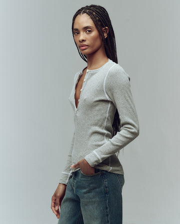 A person with long braided hair wears THE GREAT. F25 BABY RIB’s The Baby Rib Henley in Heather Grey and blue jeans, standing against a plain light background.