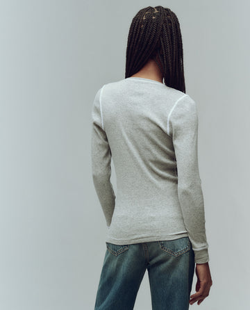 Person with long braids facing away, wearing THE GREAT. F25 BABY RIB "The Baby Rib Henley" in Heather Grey and blue jeans, against a plain background.