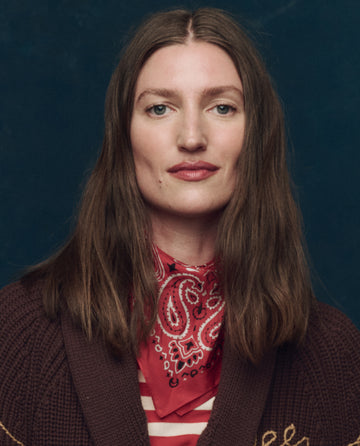 A person with long brown hair, wearing THE GREAT. F25 COLLECTORS The Silk Bandana in True Red and a brown sweater, poses in front of a dark blue background.