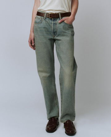 Person wearing The Wayne Jean (30" inseam) by THE GREAT. PF24 in Sedona Wash with a brown leather belt, a white 100% cotton shirt, and brown leather shoes, standing against a plain background.