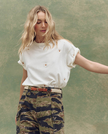 A woman with long blonde hair wears THE GREAT. SU25 The Boxy Crew. Embroidered in washed white with floating daisy embroidery and camouflage pants, standing before a mottled green backdrop.