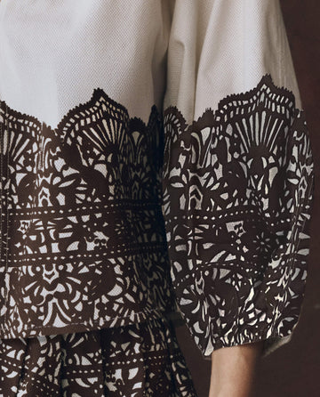 A close-up shows a person wearing THE GREAT. F25 D1 The Chariot Top in cream, featuring an intricate black western lace print across the chest and sleeves, with their arm relaxed at their side.