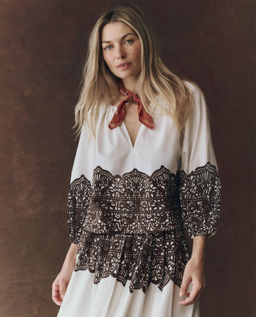 A woman with long blonde hair wears THE GREAT. F25 D1's The Chariot Top in cream with Western lace print, paired with a red neck scarf, standing against a brown background.