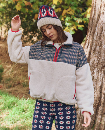 Someone stands outdoors near a tree, wearing THE GREAT. F25 TGO’s Print-Mix Plush Terrain Half-Zip in Mini Frosted Daisy & Cream, paired with a patterned knit hat and floral print pants. Green foliage fills the background.