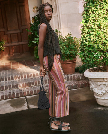 A person with long braided hair stands on a pathway next to a potted plant, wearing a sleeveless top, striped pants, and sandals, holding The Crochet Drawstring Bag in black from THE GREAT. SU23, which is hand-woven from natural Agave plant fibers.