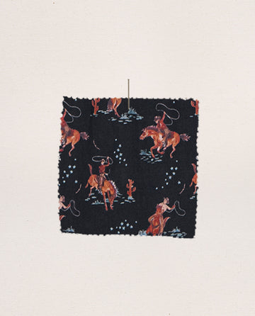 The Pajama Shirt – Navy Cowboy by THE GREAT. SP25 SLEEP features a square fabric swatch with cowboys riding horses and lassoing on a black background, accented with blue and white stars—ideal for unique sleepwear or pajama shirts.