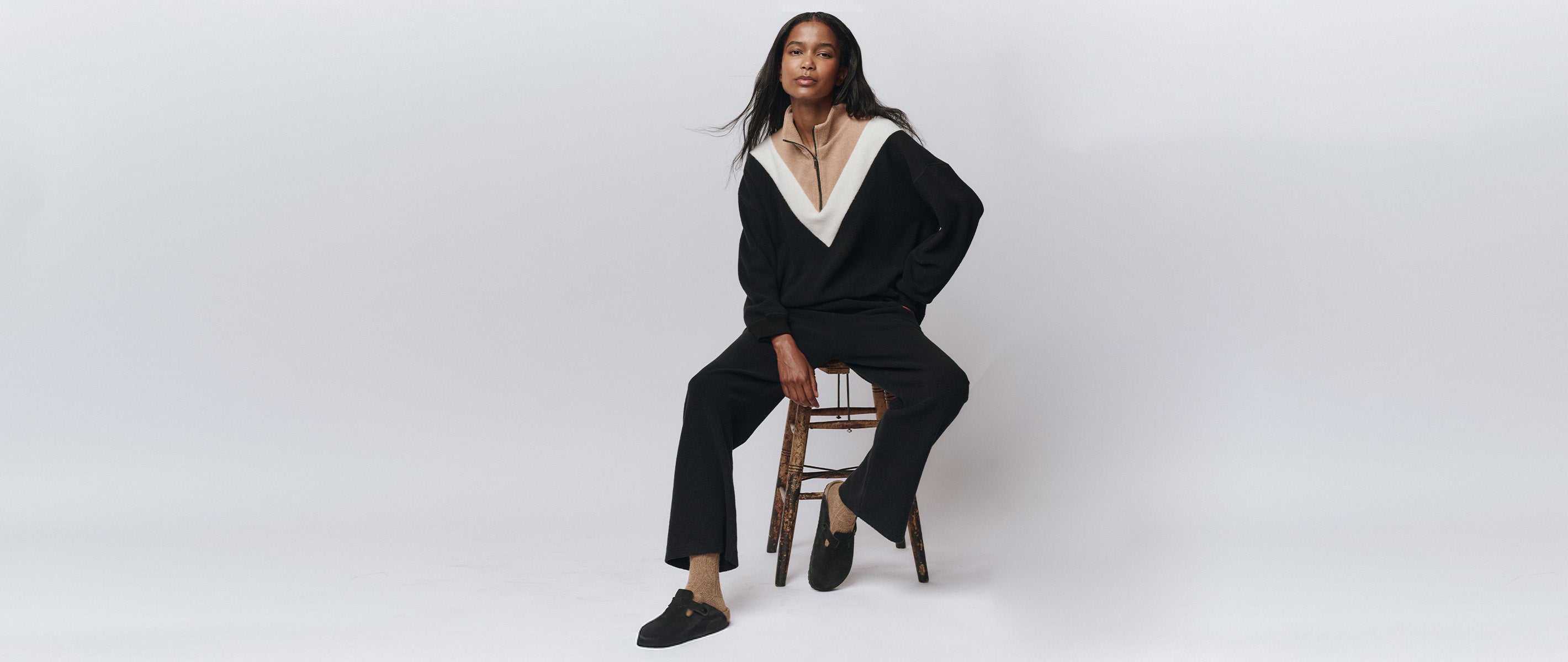 A woman sits on a wooden chair against a plain white background, wearing a black pantsuit with a white V-neck collar and black shoes.