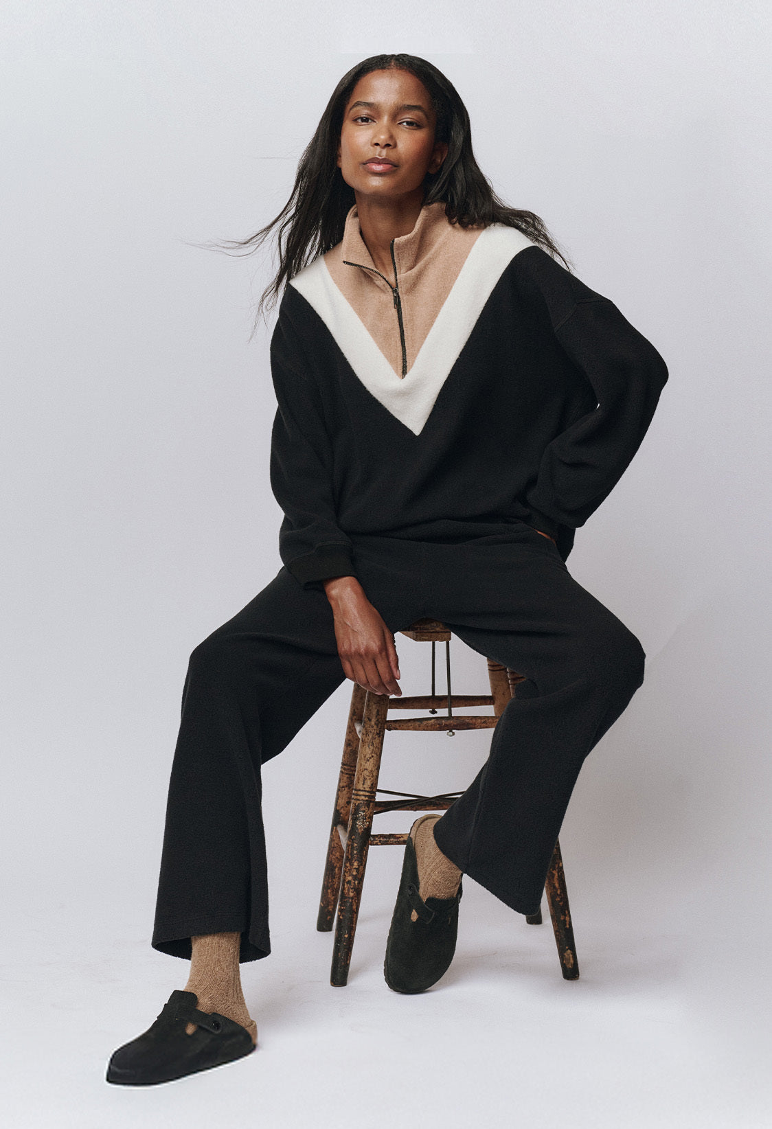 Person sitting on a wooden stool, wearing a black and beige zip-up top with a white V pattern, black pants, beige socks, and black slip-on shoes, against a plain light background.