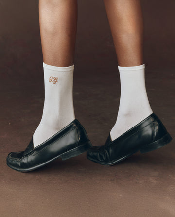 Someone wearing THE GREAT. F25 D1 ACCESSORIES' The Monogrammed Trouser Sock in Cream, featuring "TG" monogramming, and black loafers, stands on a brown surface.