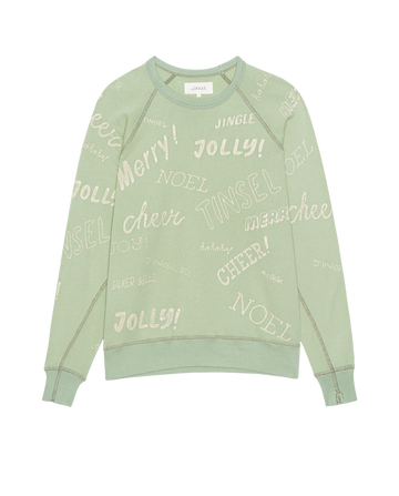 The College Sweatshirt by THE GREAT. H25 HOLIDAY GRAPHICS features a vintage mint color and festive holiday greetings like "Jolly!", "Merry!", "NOEL," "TINSEL," and "cheer" in playful fonts across the fabric.