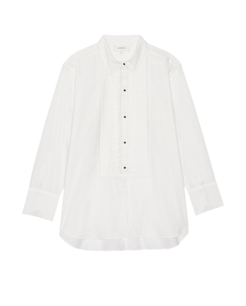 The Classic Tux Top in white by THE GREAT. H25 COLLECTION features a pointed collar, pleated front, and black buttons, adding a refined twist to the classic style, showcased against a plain background.