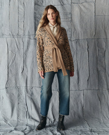 A woman stands against a gray textured backdrop, wearing THE GREAT. H25 COLLECTION’s The Bandana Riding Cardigan in Camel over a tan sweater, paired with blue jeans and black ankle boots.