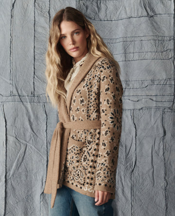 A woman with wavy blonde hair models THE GREAT. H25 COLLECTION's The Bandana Riding Cardigan in Camel, featuring black and cream patterns, over a white shirt and blue jeans against a textured gray backdrop.