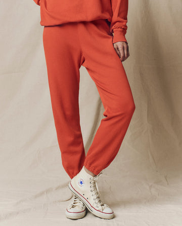 A person wearing The Stadium Sweatpant. Solid in Desert Clay by THE GREAT. SU25 SEASONAL KNITS stands with one leg crossed over the other against a neutral background, paired with white high-top Converse sneakers.