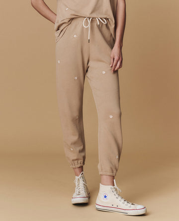 A person wears THE GREAT. PF25 EMB KNITS The Stadium Sweatpant. Embroidered in khaki with daisy embroidery, paired with a matching top and white high-top Converse sneakers, standing against a beige background.