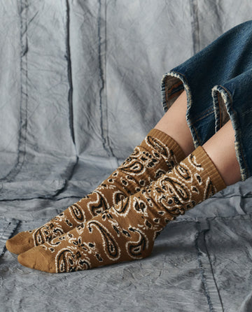 A person wearing THE GREAT. H25 COLLECTION The Bandana Sock in Camel, featuring a paisley pattern, styled with frayed hem blue jeans and seated on a textured gray surface.